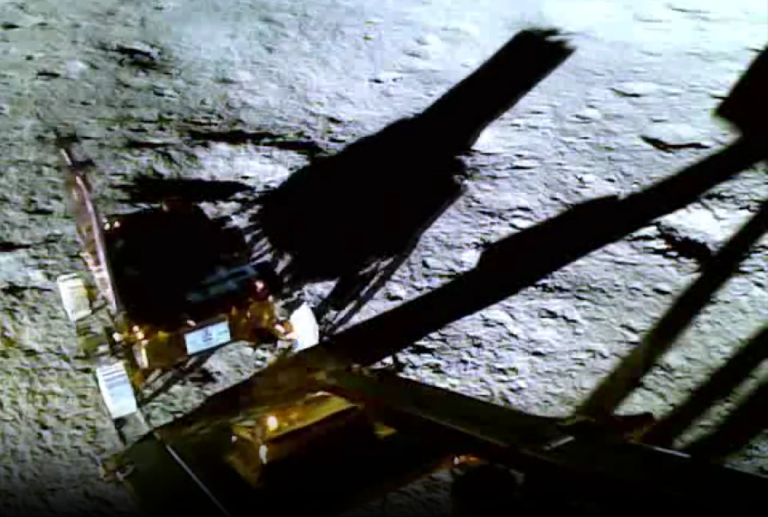 a gold colored small rover called pragyaan with white wheels travelling down a gold colored ramp of the vikram lander, on the rough silver colored surface of the moon